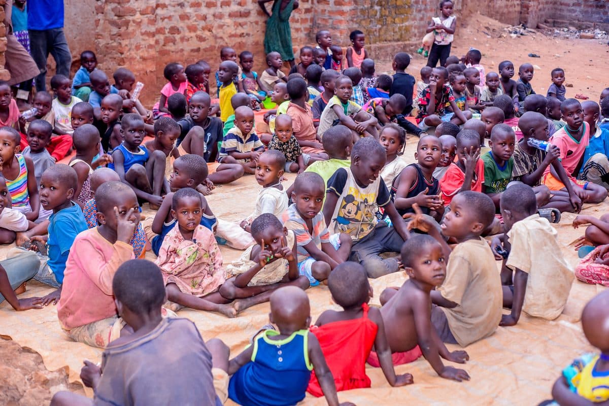 A large group of children sitting together on the ground in a courtyard, waiting for the daily hot meal to be served