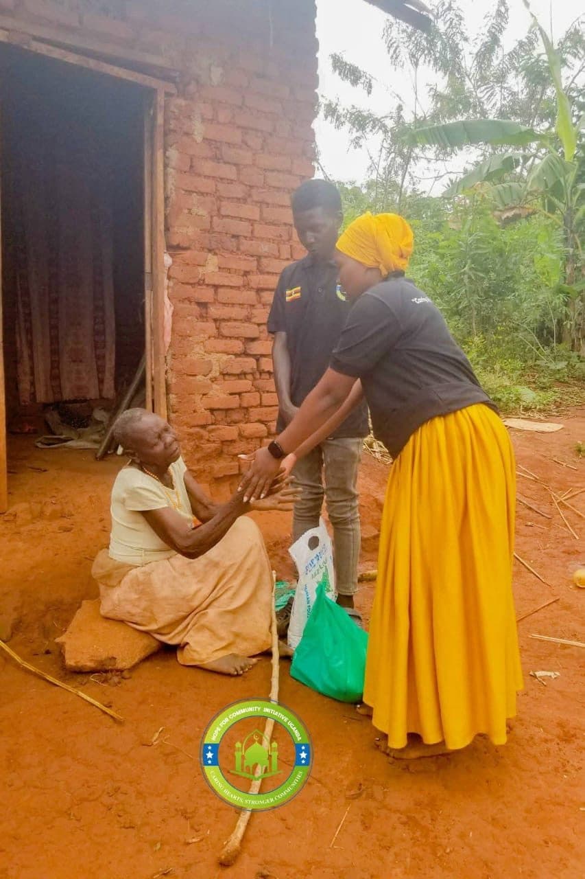 An HCIU volunteer crouching to greet an elderly woman seated outside her home, with a parcel of aid beside them